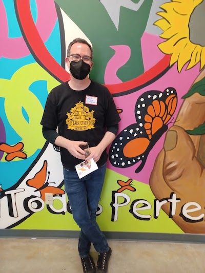 A person wearing a mask, black t-shirt with yellow writing, and jeans, stands in front of a wall with brightly colored murals, including a large monarch butterfly and some smaller butterflies.