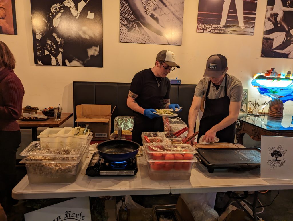 Members of Iron Roots Cooperative stand behind a table. On the table is food, a frying pan, and a griddle. They are putting food on a plate.